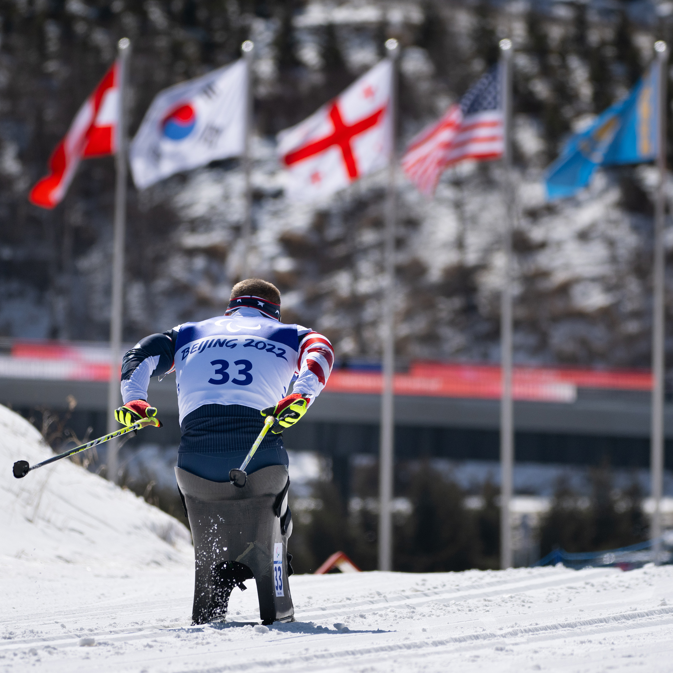 Dan Cnossen competes in skiing event, racing over snow with flags and trees in the distance