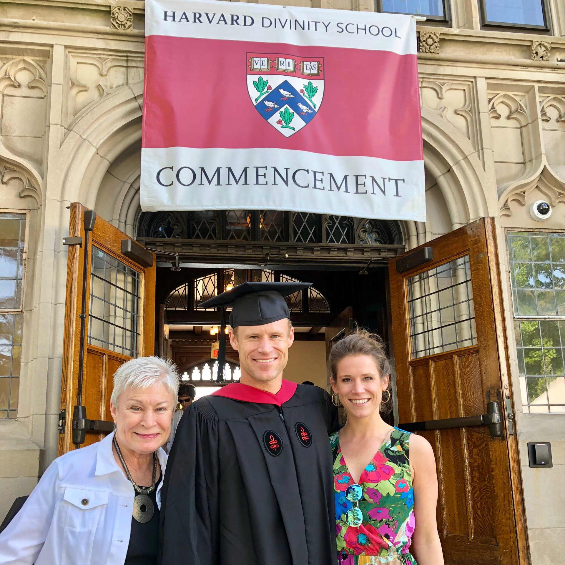 Dan Cnossen with family members in front of Swartz Hall during commencement. A Harvard Divinity School Commencement flag is above the three people