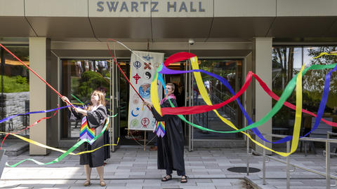 graduating students in gowns wave ribbons in front of a campus building ahead of commencement