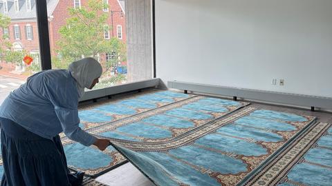 person preparing for religious service by laying out light blue prayer rugs in room with window to left