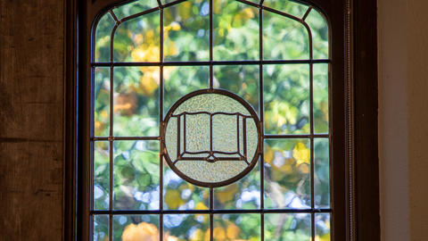 Swartz Stained Glass Window with centerpiece displaying an open book.  Fall tree leaves are visible beyond the window.