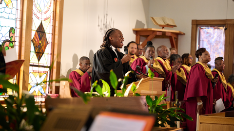 A student stands in front of a church preaching with a choir behind. They are all wearing choral robes