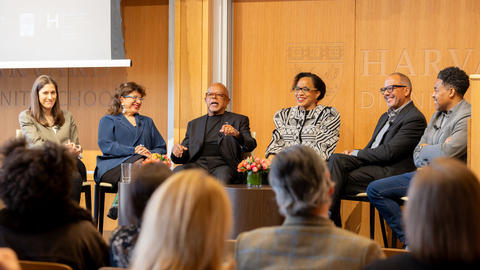 six people sit next to each other in a row speaking together and smiling with a brown background behind and audience members in front