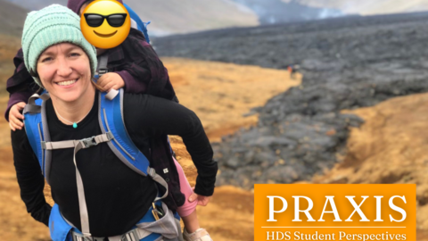 HDS Student Amy Sexauer stands at the top of a mountain with her child on her back. The child has a smiley face emoji over their face.