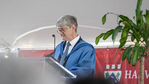 Dean David Hempton standing behind a clear podium dressed in a light blue academic robe