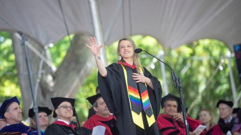 Lindsay Sanwald in black academic regalia holding her right hand up standing in front of seated people during Harvard's Commencement ceremony