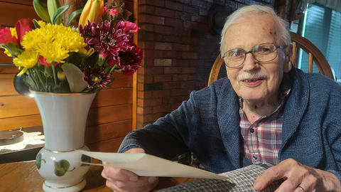 HDS Professor of Ethics Emeritus Arthur Dyck seated at a table holding a piece of paper in his hand next to a vase containing flowers of various colors.