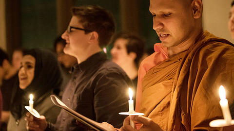Bhante Kusala holding a lit candle looking down at a piece of paper in a crowd of people holding candles in Williams Chapel