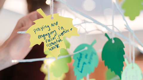 A tight shot of a wire tree with paper leaves. The yellow leave in focus reads "praying and engaging in racial justice and restorative work"
