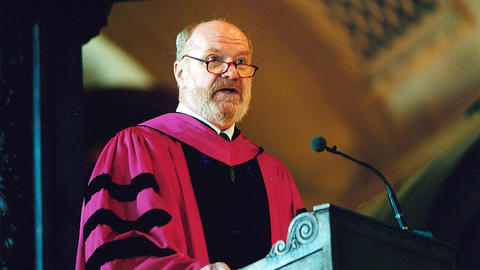 Paul Hanson in a red academic robe speaking at a podium