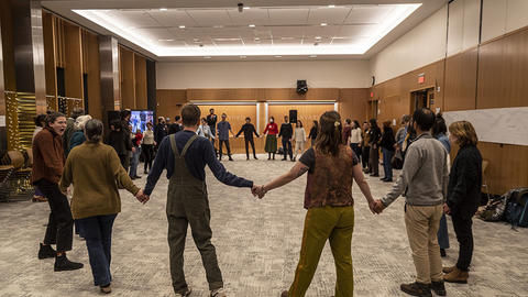 HDS students, staff, and guests join in a contra dance. The dancers hold hands in a large circle; the space in the center of the circle is empty.