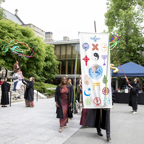 The HDS class of 2026 processes toward the Campus Green behind a multireligious banner and waving ribbons