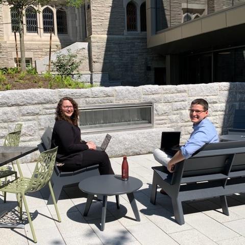 Two people sitting outside looking at the camera near entrance to Swartz Hall