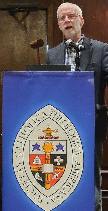 HDS Professor Francis X. Clooney, S.J., stands holding a gavel behind a podium with the logo of the Catholic Theological Society of America.