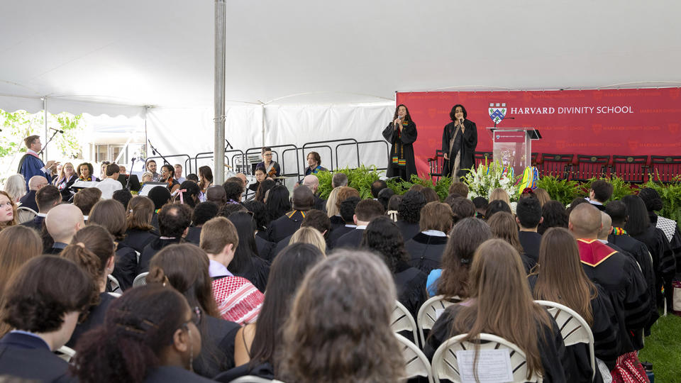 Two graduating students dressed in graduation robes sing side by side