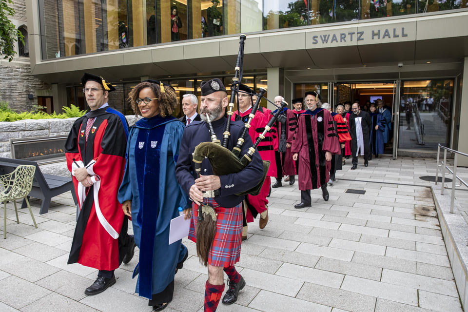 faculty adorned by academic gowns walk in a line with a building behind them
