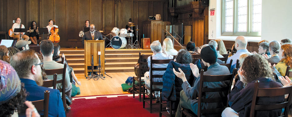 people seated in chapel looking toward stage and lectern with people playing music and speaking