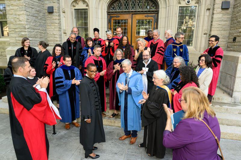 President Claudine Gay stands outside Swartz Hall in the middle of a group of HDS faculty members all wearing academic regalia who applaud President Gay