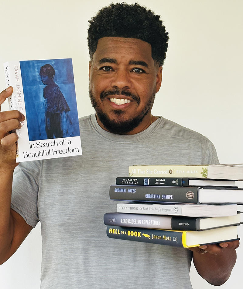 Terrence L. Johnson stands holding a pile of stacked books in one hand and a single book upright in the other hand