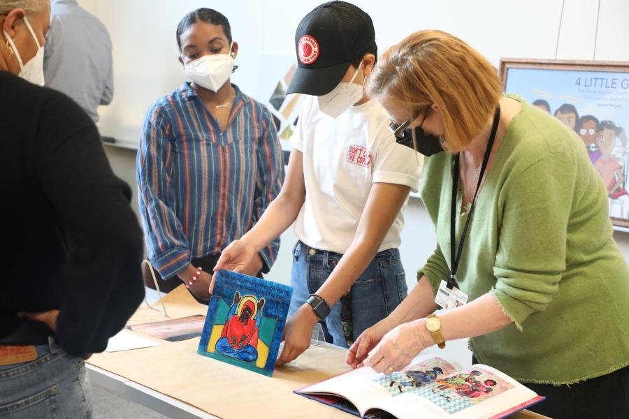 A student in a white T-shirt and dark baseball cap holds up a work of stained glass next to students and a library staff member.