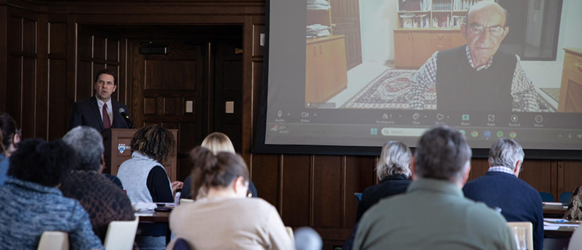 man at left speaks from the podium to a virtual guest who is seen on screen at center