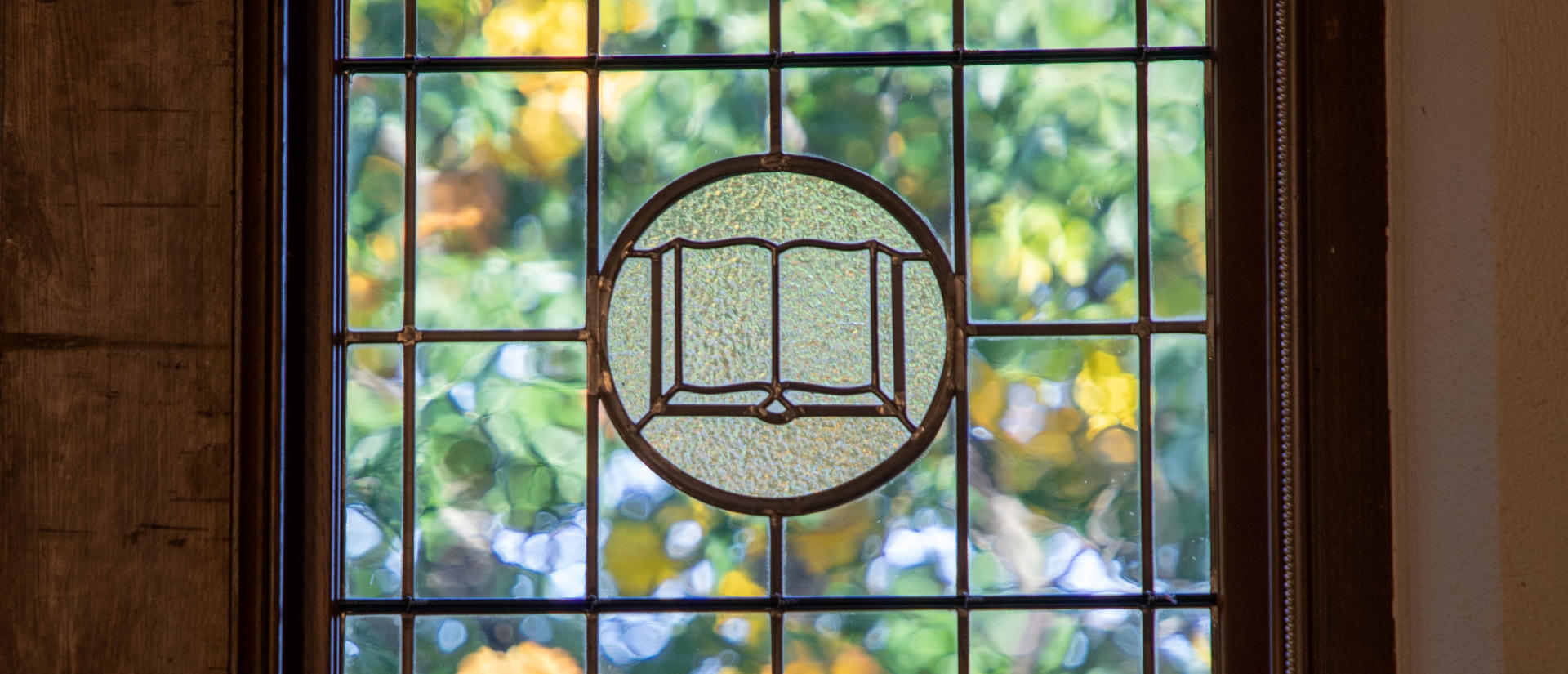 Swartz Stained Glass Window with centerpiece displaying an open book.  Fall tree leaves are visible beyond the window.