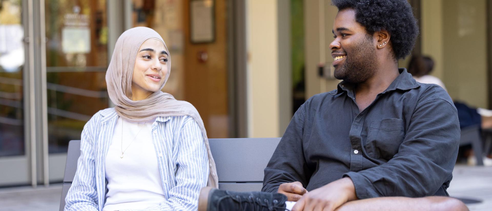 Two HDS students sit next to each other and smile, mid-conversation, in front of the main entrance to Swartz Hall.  
