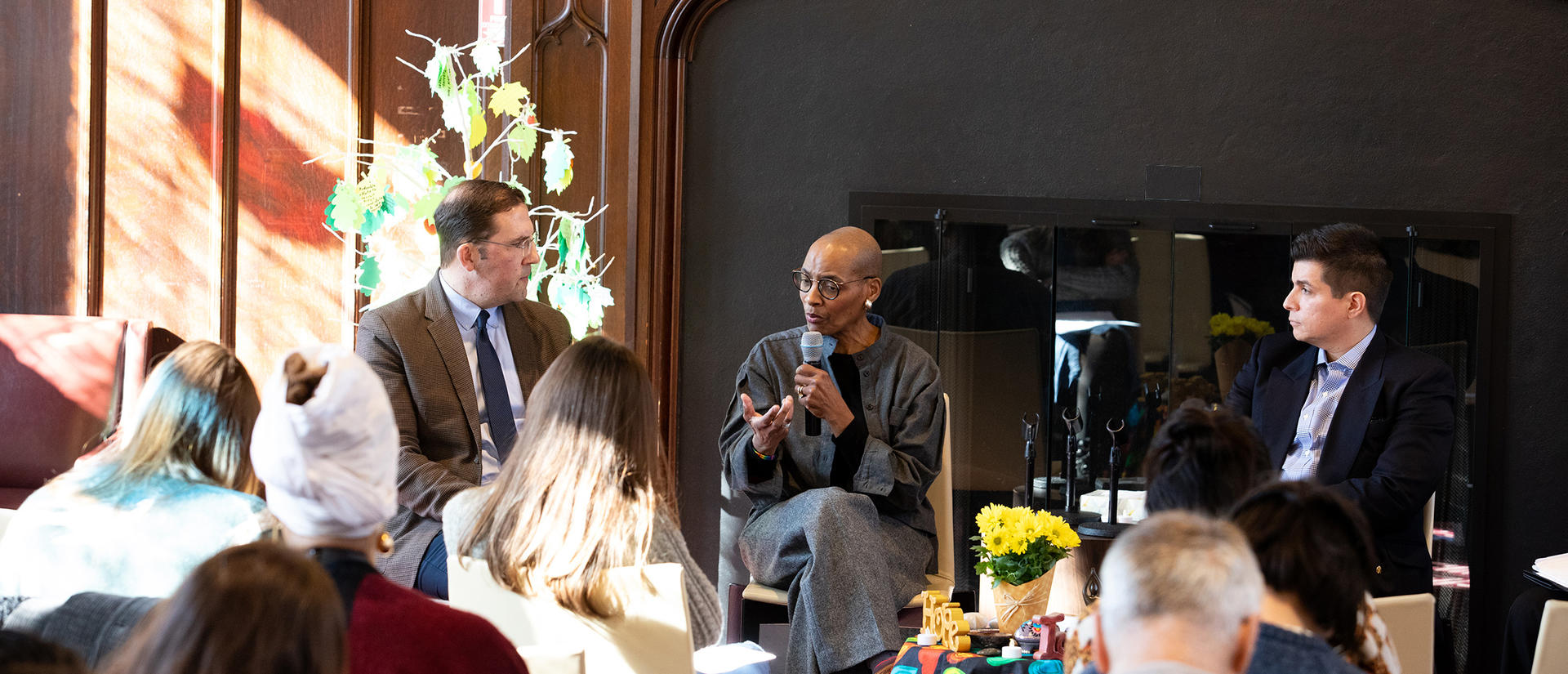 David Holland, Kelly Brown Douglas, and Raul Zegarra sit side-by-side during a conversation, with brown wall behind them and sunlight coming in on the left side