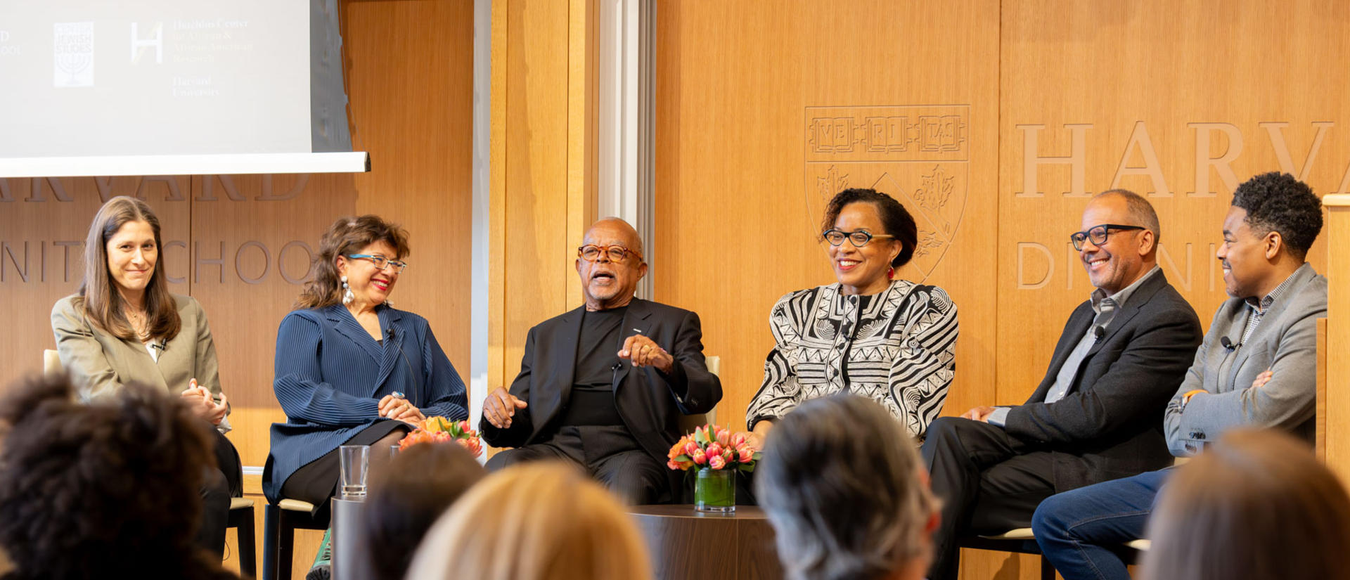 six people sit next to each other in a row speaking together and smiling with a brown background behind and audience members in front