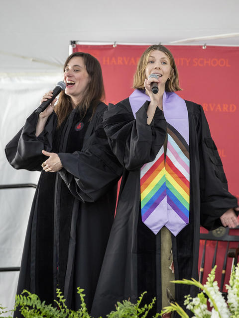 Two graduating students dressed in graduation robes sing side by side