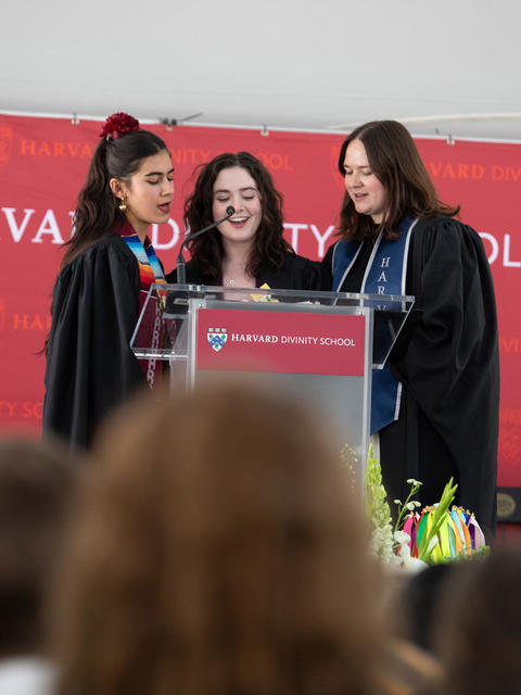 three graduating students recite a prayer during a commencement service in front of a red backdrop