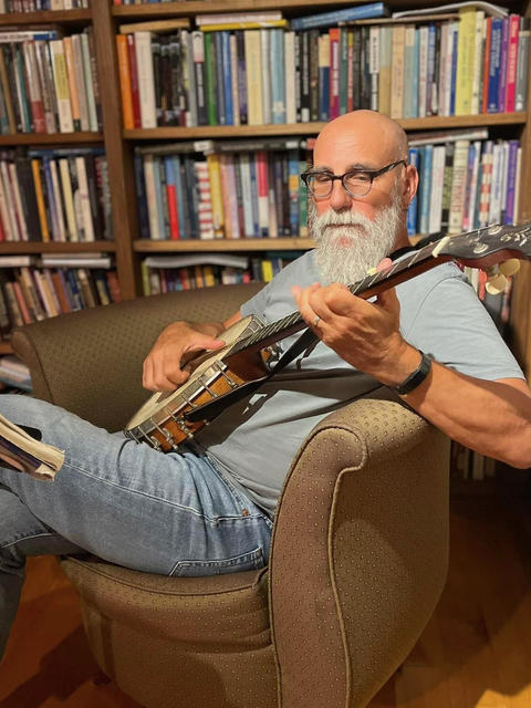 Shaul Magid playing banjo sitting down in a chair, surrounded by books