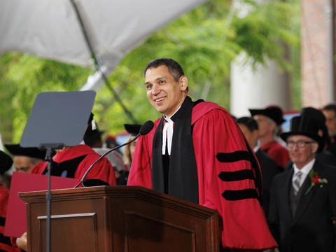 a person in a formal academic gown smiles and delivers remarks at a podium in front of persons seated in gowns